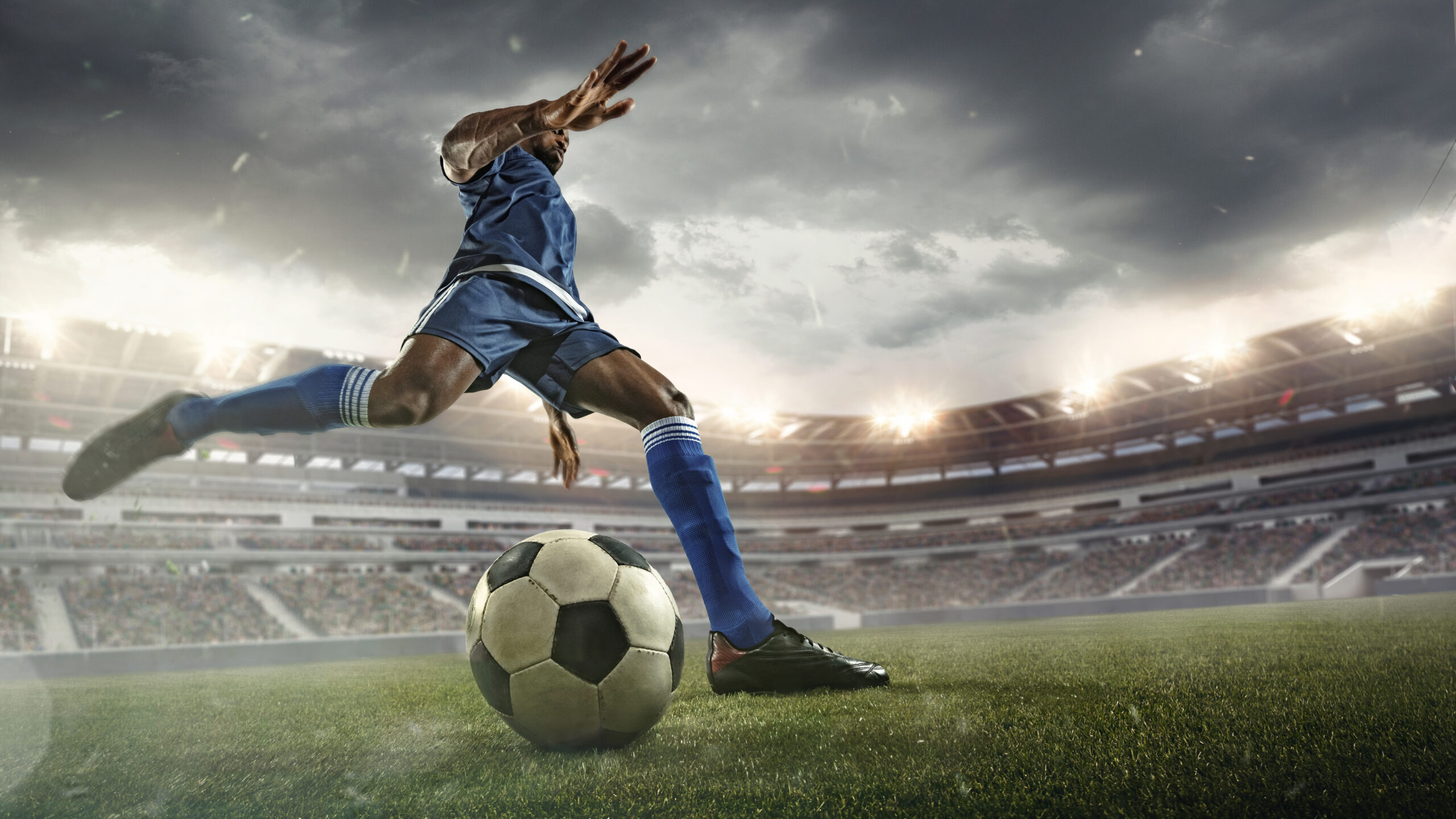 A footballer in a blue shirt kicking a ball in a football field.