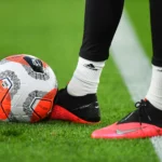 A picture of a football player's boots besides a ball on a football field.